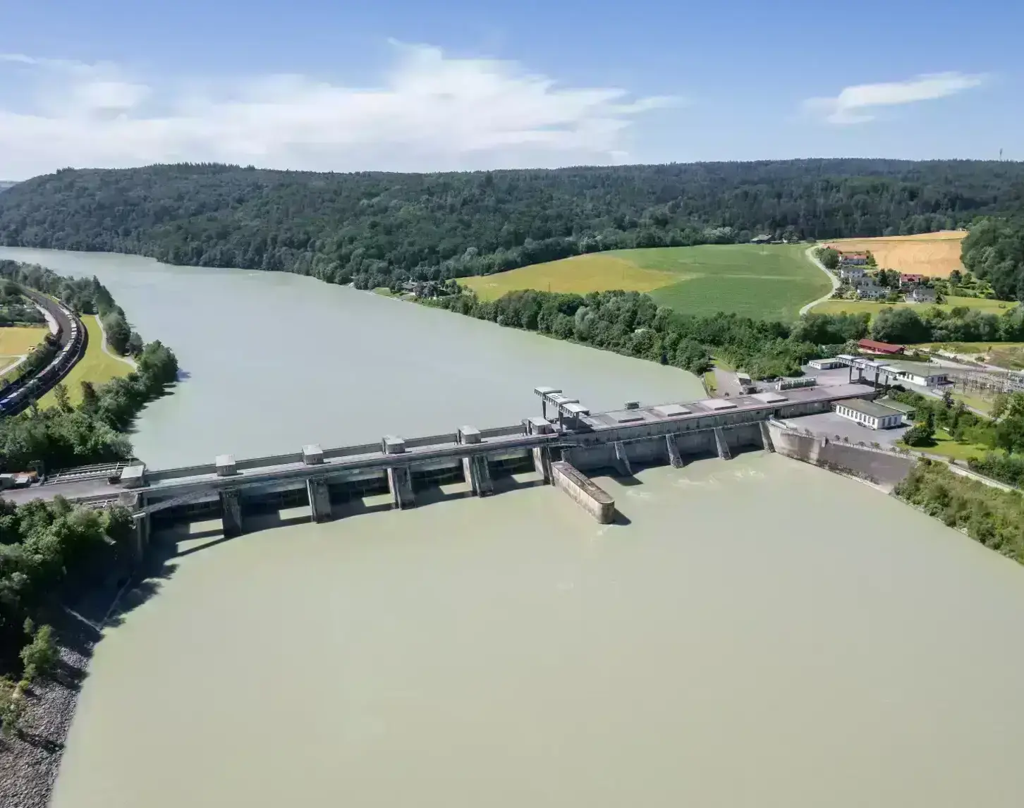 Das Bild zeigt eine Luftaufnahme des Kraftwerks Passau-Inglings von der Seite flussabwärts. Der Inn ist dabei blaugrün und hebt sich dadurch stark von den hell- und dunkelgrünen Wiesen und Wädern links und rechts des Flusses ab. Im Hintergrund sieht man noch den Verlauf des Inns, dahinter Wald sowie blauen Himmel.