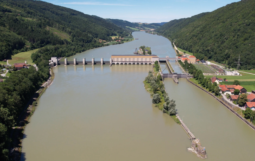 The Jochenstein power station can be seen from the air on a sunny day. While the Danube is coloured green-blue, the trees on the banks to the left and right are a lush dark green. While the weir fields can be seen on the left, the lock for shipping can be seen on the right.  In the background you can still see the landscape and the course of the Danube upstream.