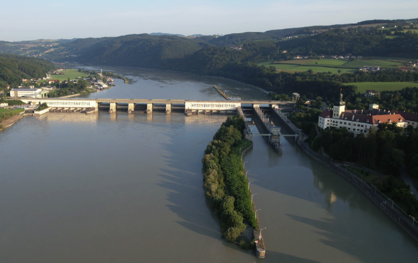 Auf dem Bild ist das Donaukraftwerk Ybbs-Persenbeug aus der Luft von der Flussabwärtsseite sichtbar. Während man im Hintergrund die schöne Landschaft mit vielen Wäldern und dem Verlauf der dunkelblauen Donau sieht, erstrahlt rechts der Ort Persenbeug im Sonnenschein. Im Hintergrund sieht man noch den blauen Himmel oberhalb der Landschaft.