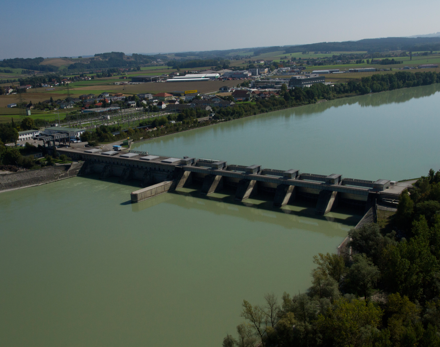 Das Bild zeigt eine Luftaufnahme des Wasserkraftwerks Schärding-Neuhaus. Im Mittelpunkt ist das Kraftwerk und die blaugrüne Donau zu sehen. Im rechten Bildrand sieht man Wald und hinter dem Kraftwerk ist die Stadt Schärding sowie die Landschaft mit strahlend blauen Himmel sichtbar.