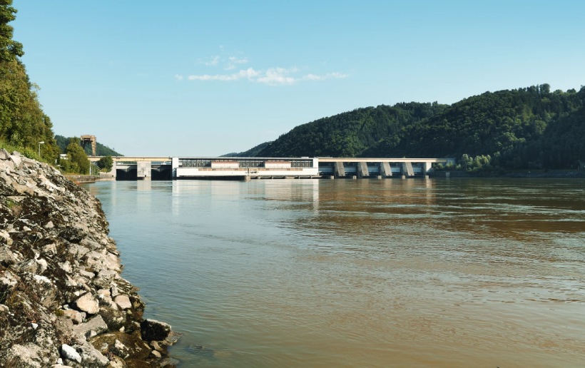 The Aschach power station can be seen from the downstream side on a beautiful summer's day. On the left you can see rocks and green trees above. In front of the power station you can see the greenish Danube flowing downstream.  To the right of the power station you can see dark green forests and behind the power station you can see a bright blue sky.