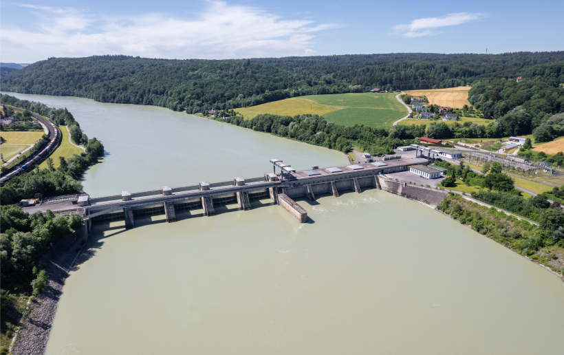 Das Bild zeigt eine Luftaufnahme des Kraftwerks Passau-Inglings von der Seite flussabwärts. Der Inn ist dabei blaugrün und hebt sich dadurch stark von den hell- und dunkelgrünen Wiesen und Wädern links und rechts des Flusses ab. Im Hintergrund sieht man noch den Verlauf des Inns, dahinter Wald sowie blauen Himmel.