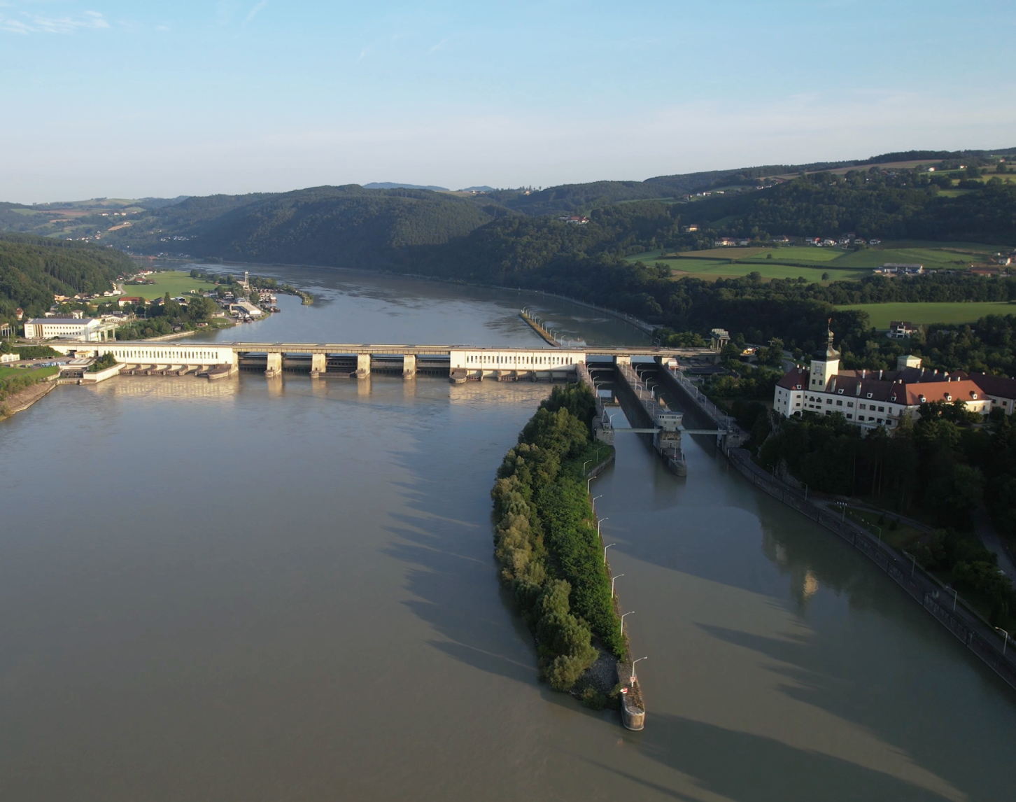 In the picture, the Ybbs-Persenbeug Danube power station is visible from the air from the downstream side. While the beautiful landscape with many forests and the course of the dark blue Danube can be seen in the background, the village of Persenbeug shines in the sunshine on the right. In the background you can still see the blue sky above the landscape.