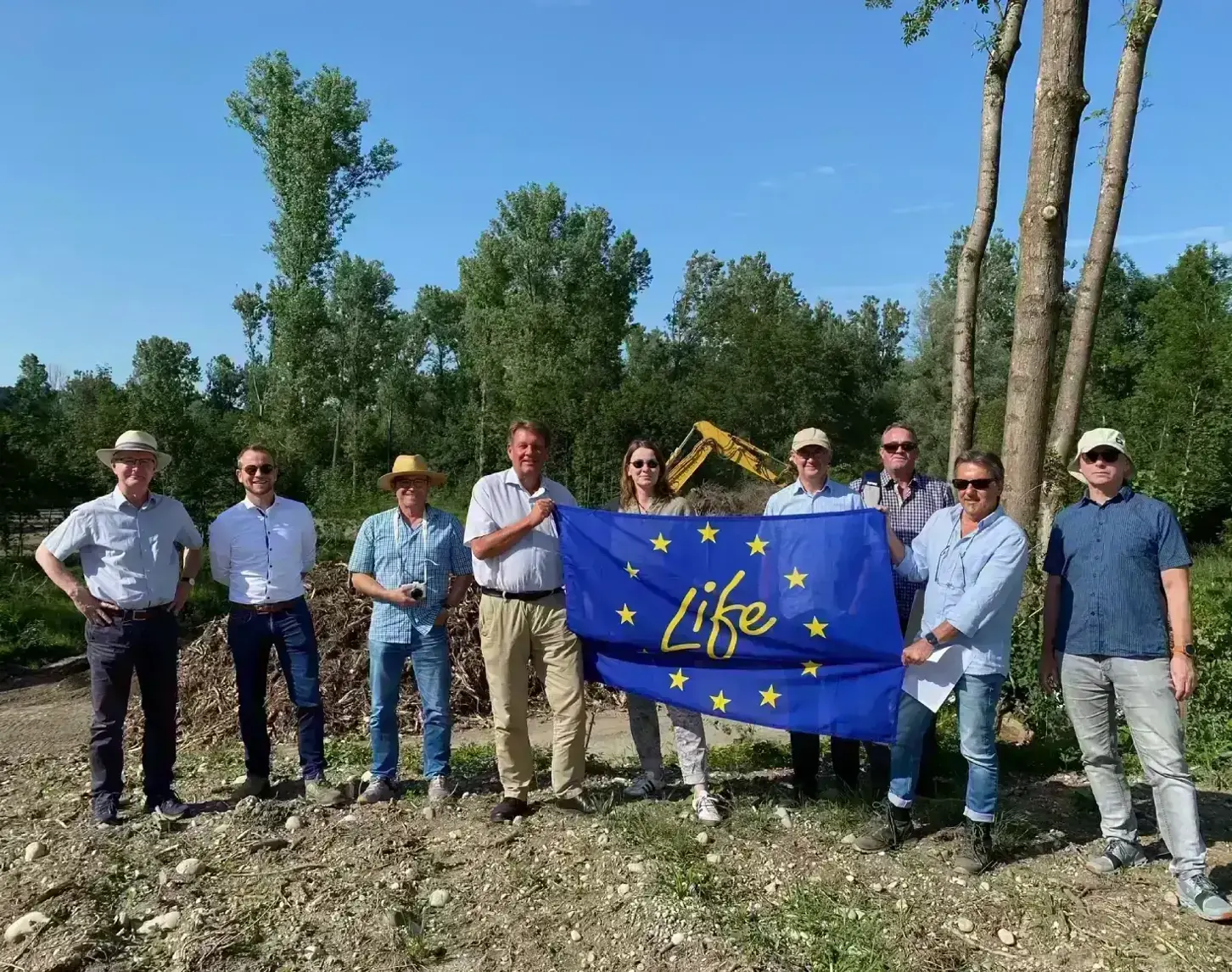 The NEEMO team poses together in the LIFE Blue Belt Danube Inn area. A large flag with the LIFE logo is held up in the centre. In the background is a bright green forest with a bright blue sky above.